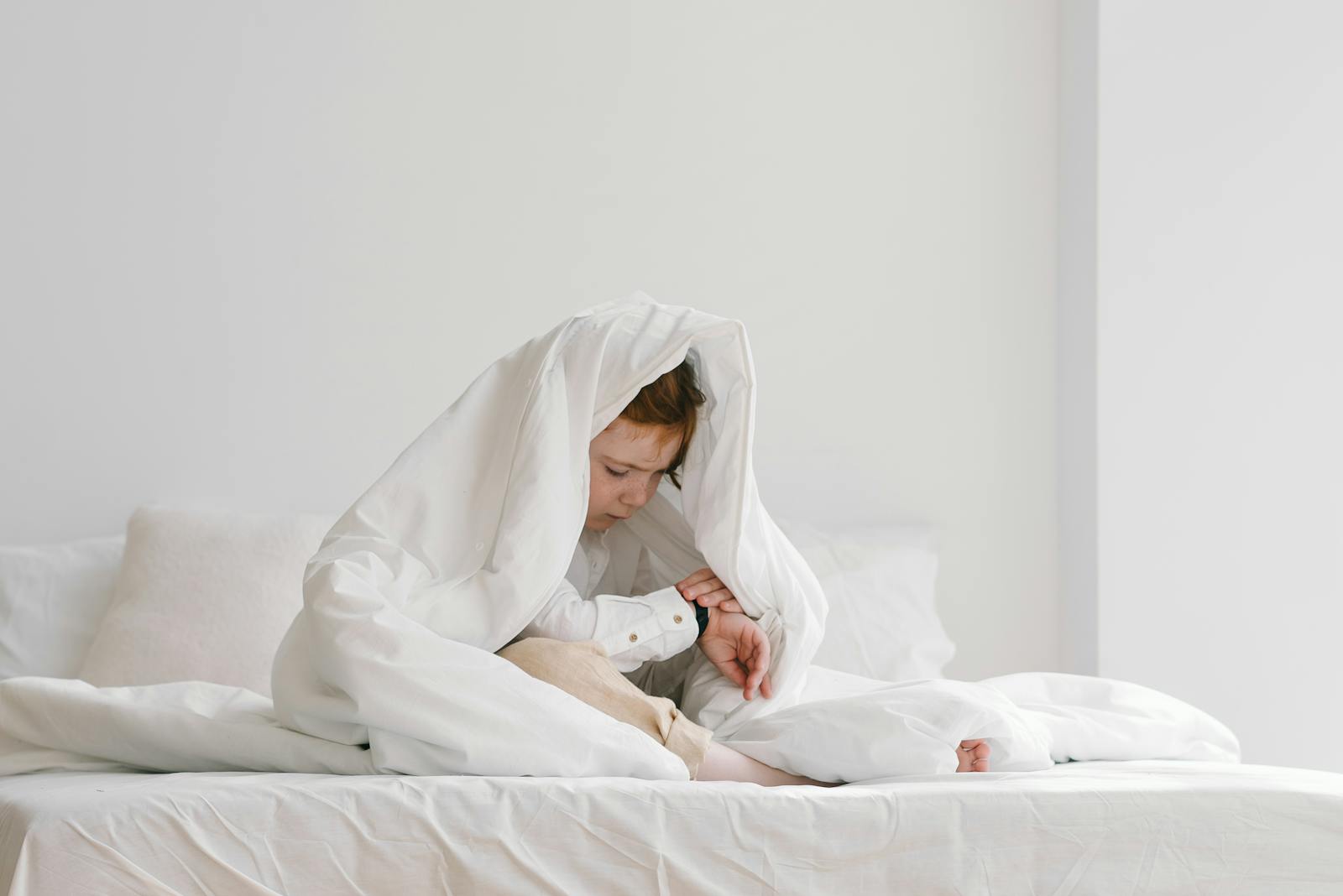 A young child sitting playfully under a white duvet on a bed in a bright room.